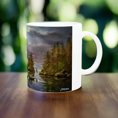 A white ceramic mug from Ken Skoda Studio, branded as "Afternoon Orca," sits on a wooden table, featuring a serene landscape painting of a forest by a lake under a cloudy sky. Birds glide over the water, and the artwork is signed "Skoda." The backdrop is a blurred mix of green and yellow, enhancing the tranquility reminiscent of an ocean scene.